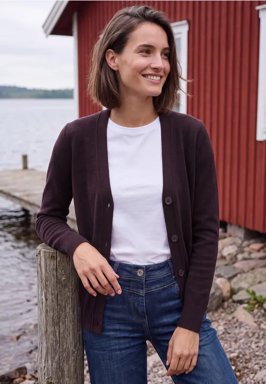 Frau in Jeans, weißem T-Shirt und violetter Strickjacke lächelt an einem Steg vor roter Holzscheune und Wasser.