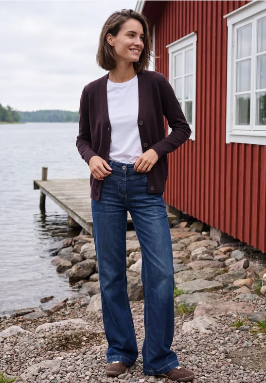 Frau steht in Jeans und Cardigan vor rotem Holzhaus am Seeufer.