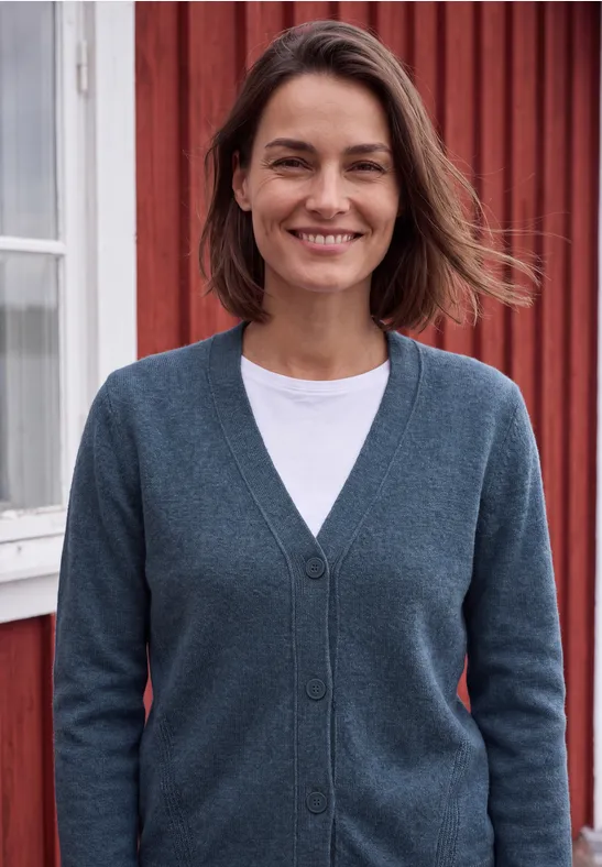 Eine Frau mit kurzem braunem Haar lächelt in einem grauen Cardigan vor einem roten Holzhaus.