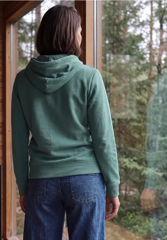 Person steht mit Blick aus dem Fenster in grüner Kapuzenjacke und Jeans, Wald draußen.