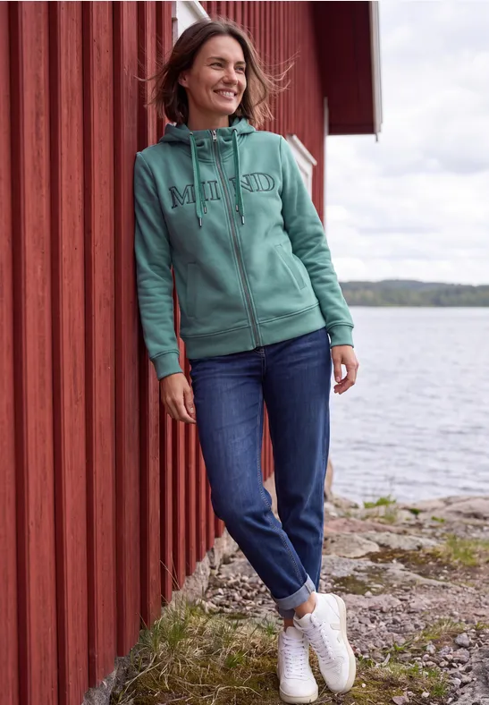 Eine Frau lehnt an einer roten Holzwand am Seeufer, trägt eine türkisgrüne Kapuzenjacke und Jeans und lächelt.