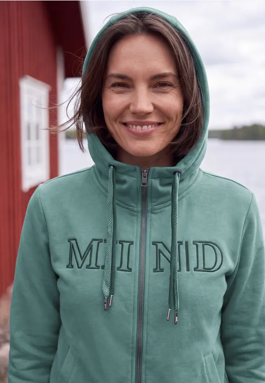 Eine lächelnde Frau steht vor rotem Haus und Wasser, trägt einen grünen Kapuzenpullover mit 'MIND'-Logo.