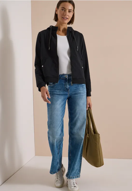Eine Frau in schwarzer Jacke, weißem Top und blauen Jeans hält eine olivgrüne Tasche.