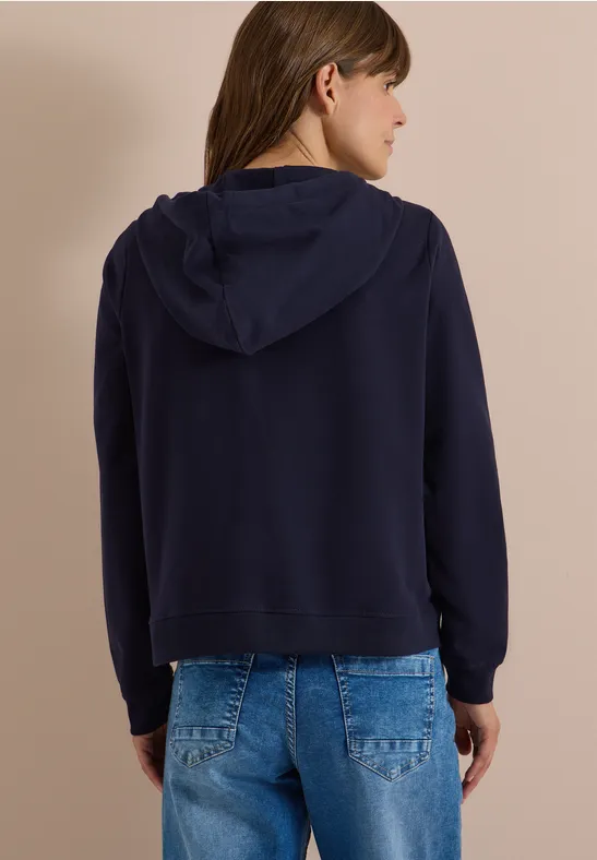 Rückansicht einer Frau mit dunkelblauer Kapuzenjacke und blauen Jeans vor beigem Hintergrund