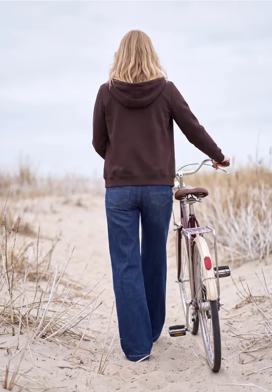 Eine Person im Kapuzenpullover schiebt ein Fahrrad über sandige Dünen am Strand.