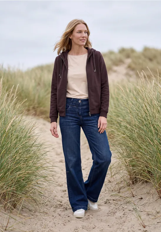 Eine Frau spaziert lächelnd in Jeans und Kapuzenjacke über sandige Dünen