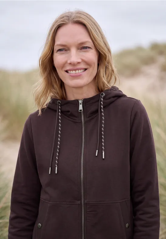 Eine lächelnde Frau in brauner Kapuzenjacke steht vor einer Dünenlandschaft.