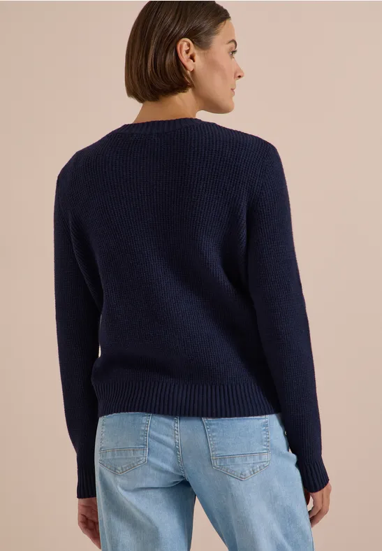 Femme vue de dos portant un pull bleu foncé et un jean bleu clair sur fond clair.