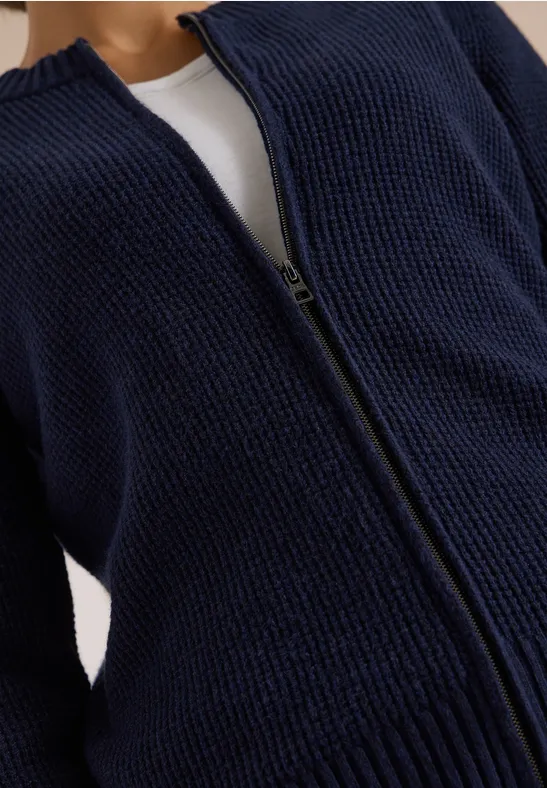 Gilet zippé bleu marine en maille épaisse, ouvert sur un haut blanc visible en dessous.