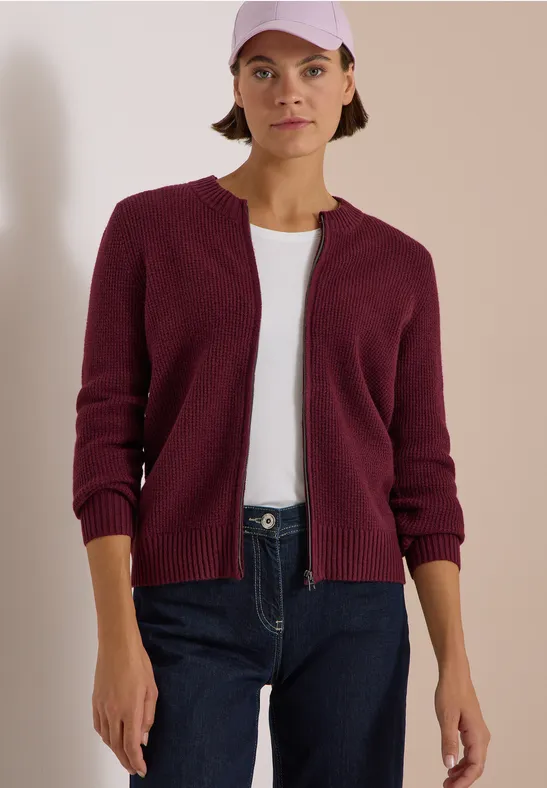 Femme en jean, t-shirt blanc, gilet bordeaux et casquette devant un fond clair.