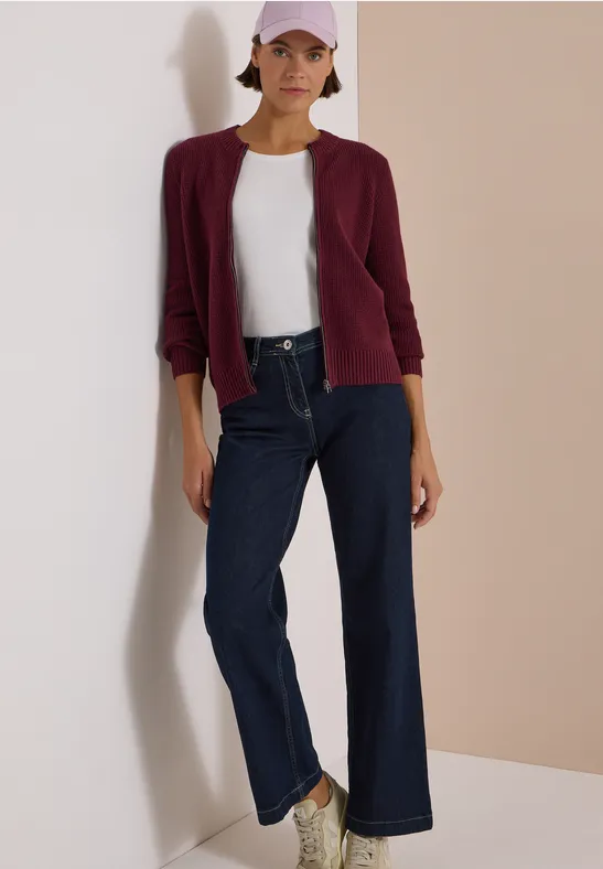 Femme en t-shirt blanc, blazer bordeaux, jean large et casquette rose devant un fond sobre.