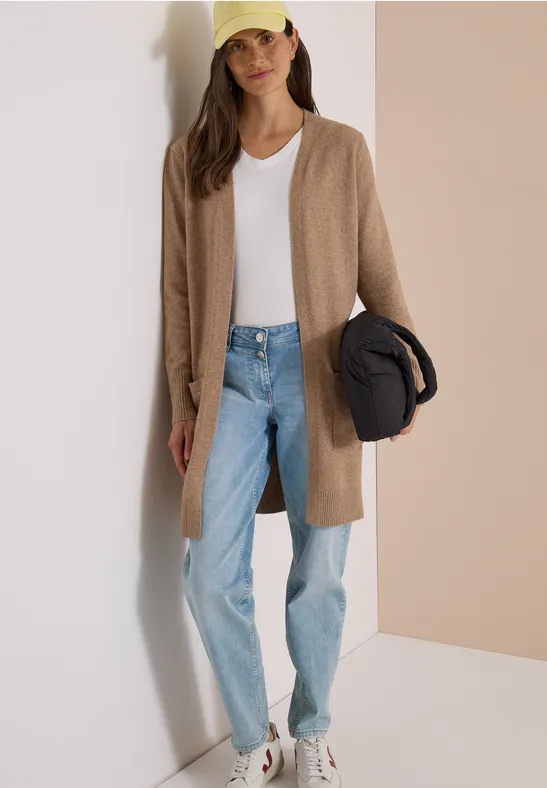 Vrouw in lange beige cardigan, wit T-shirt, lichtblauwe jeans, witte sneakers, houdt gele pet en zwarte jas vast