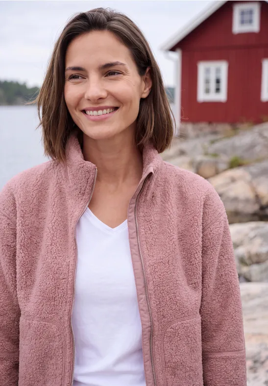 Eine lächelnde Frau in rosa Fleecejacke vor einem roten Haus am Wasser.