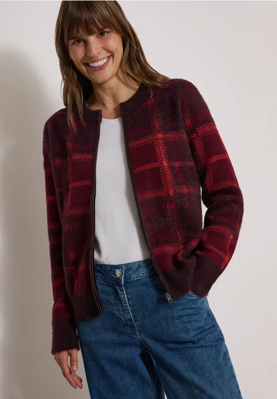 Frau lächelt, trägt karierte rote Strickjacke, weißes T-Shirt und blaue Jeans.