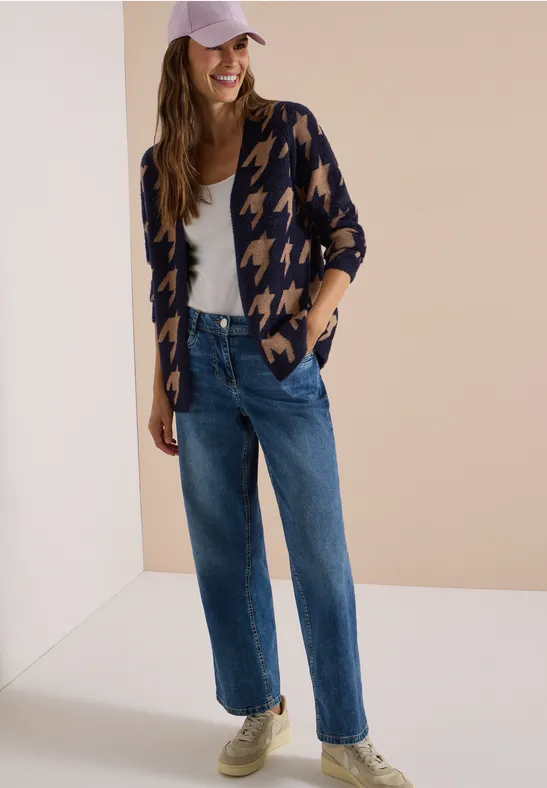Femme en jean droit, top blanc, gilet pied-de-poule et casquette, souriante devant un fond neutre.