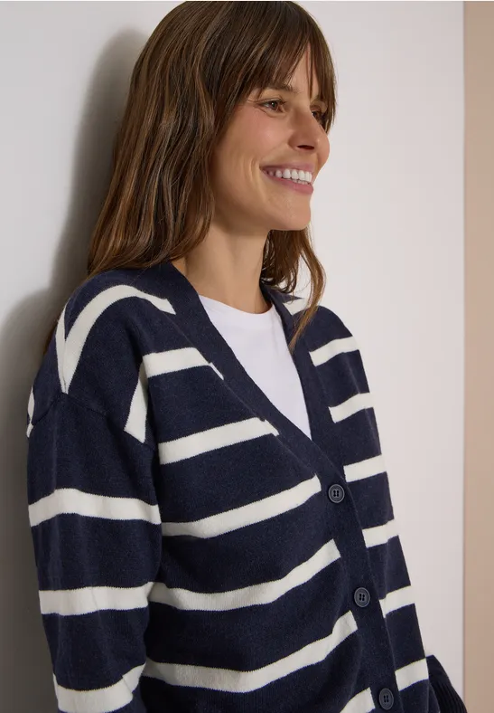 Frau lächelt und trägt einen marineblauen Cardigan mit weißen Streifen und ein weißes Shirt.