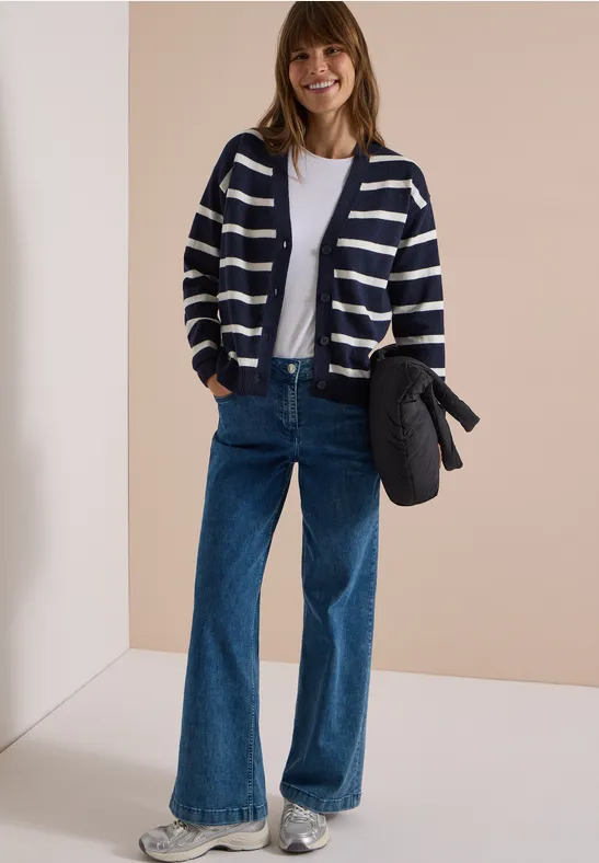 Frau in gestreifter Strickjacke, weißem T-Shirt und weitem Jeans, hält eine Tasche, lächelnd.