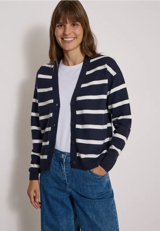 Eine lächelnde Frau trägt eine blau-weiße gestreifte Strickjacke, weißes Shirt und blaue Jeans.