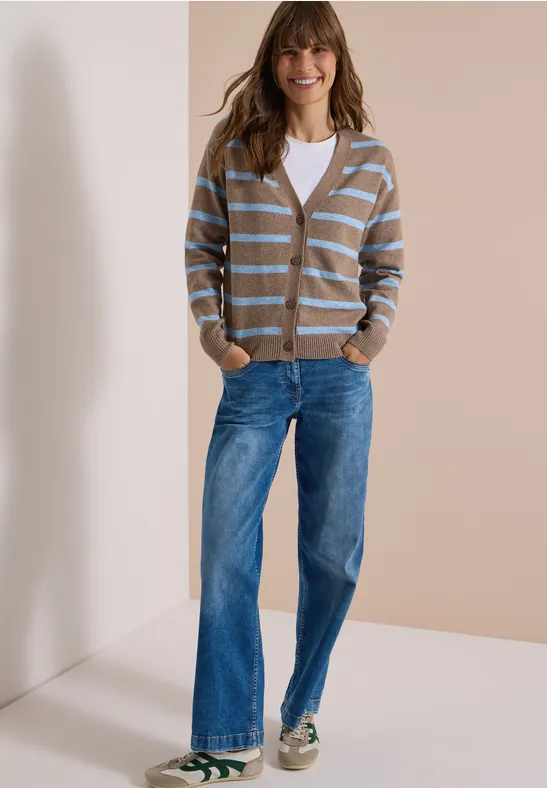 Frau in weißem Top, gestreifter Strickjacke, weiten Jeans und Sneakern vor neutralem Hintergrund.