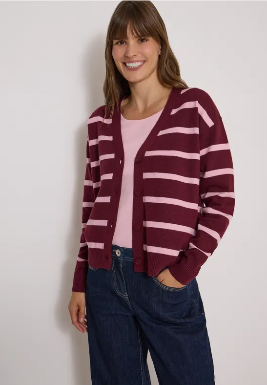 Eine lächelnde Frau in bordeauxroter, gestreifter Strickjacke, rosa Top und jeansblauer Hose.
