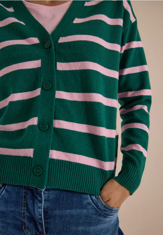 Person trägt eine grün-rosa gestreifte Strickjacke und Jeans, eine Hand steckt in der Hosentasche.