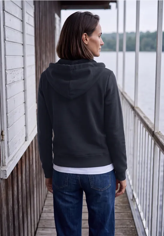 Frau in schwarzer Kapuzenjacke und Jeans steht auf hölzernem Steg und blickt aufs Wasser.
