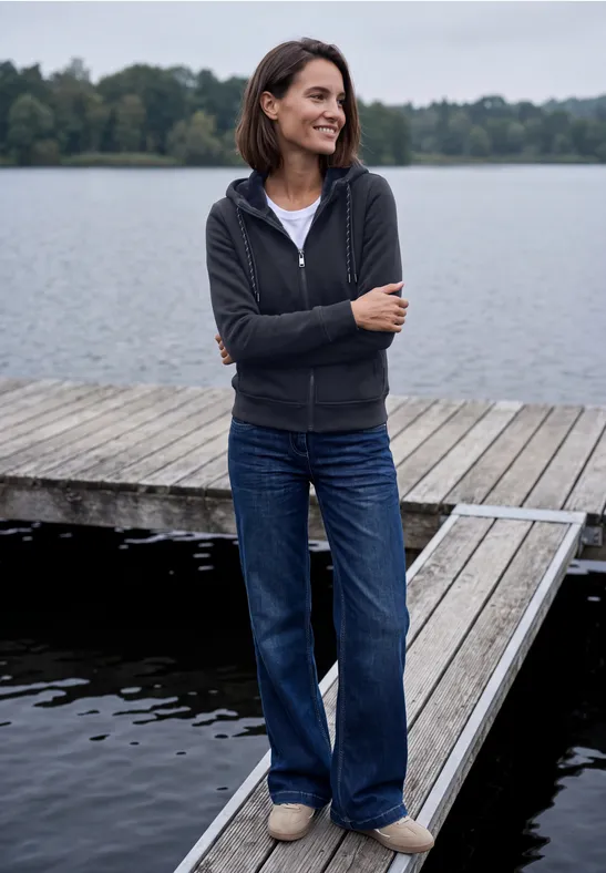Eine Frau steht lächelnd am Ende eines Holzstegs am See, trägt Jeans und eine dunkle Kapuzenjacke.