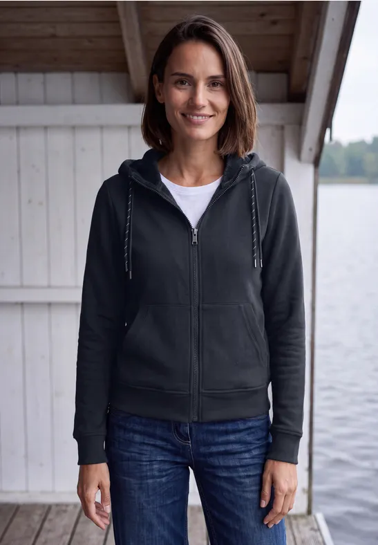 Eine Frau steht lächelnd vor einer Seehütte, trägt Kapuzenjacke, T-Shirt und Jeans.