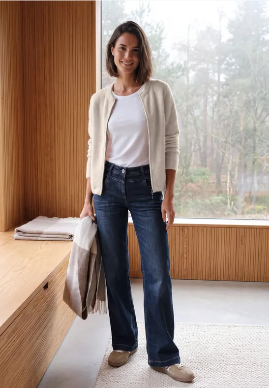 Vrouw in wit T-shirt, licht vest, spijkerbroek en schoenen, met jas in hand in een moderne ruimte.