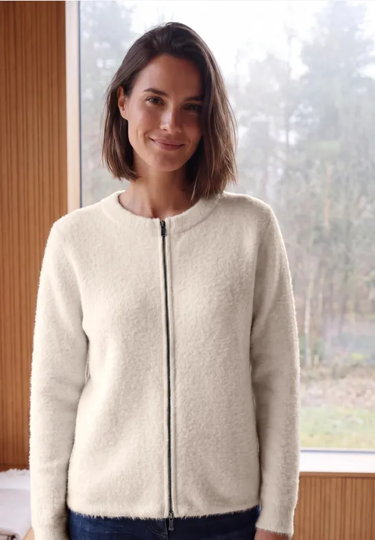 Een vrouw draagt een crèmekleurige teddyfleecejas met rits, staand voor een raam met boszicht.