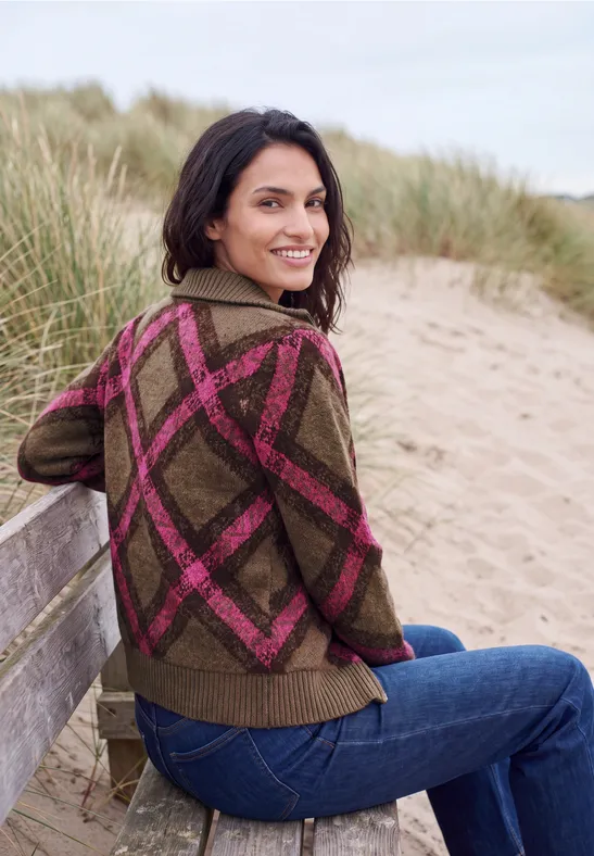 Eine Frau sitzt lächelnd auf einer Strandbank, trägt eine grüne Jacke mit pinken Rautenmuster und Jeans.