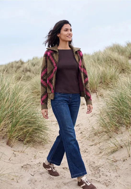 Frau in Musterjacke und Jeans spaziert lächelnd über sandige Dünen