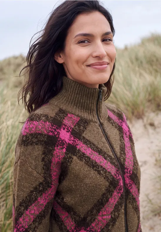 Frau am Strand in einem braunen Wollpullover mit pinkem Karomuster und Reißverschluss.