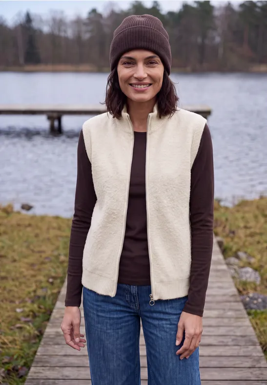 Frau mit braunem Pullover, beiger Fleeceweste, Jeans und Mütze steht auf Holzsteg am See.