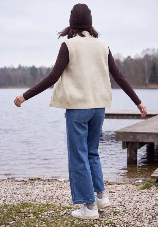 Person in Jeans, weißer Weste und Mütze spaziert am Seeufer neben einem Holzsteg.