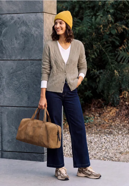 Frau in gelber Mütze, grauem Cardigan und blauer Hose mit Reisetasche vor modernem Gebäude und Bäumen.