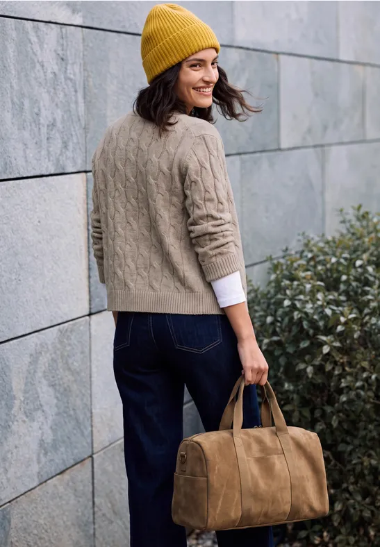 Eine Frau mit gelber Mütze und grauem Pullover trägt lächelnd eine braune Reisetasche an einer Steinmauer.