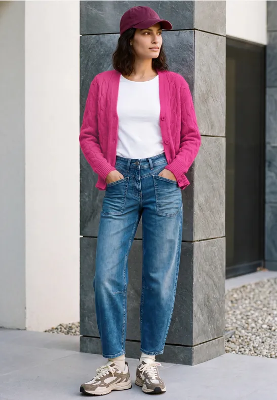 Eine Frau in pinkem Cardigan, weißem Shirt, Jeans, Sneakern und Cap steht vor moderner Fassade.