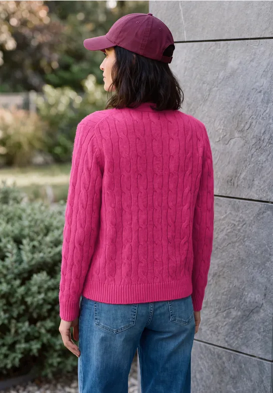 Frau mit pinkem Strickpullover, blauen Jeans und roter Cap steht rückwärts vor einer grauen Wand im Freien.