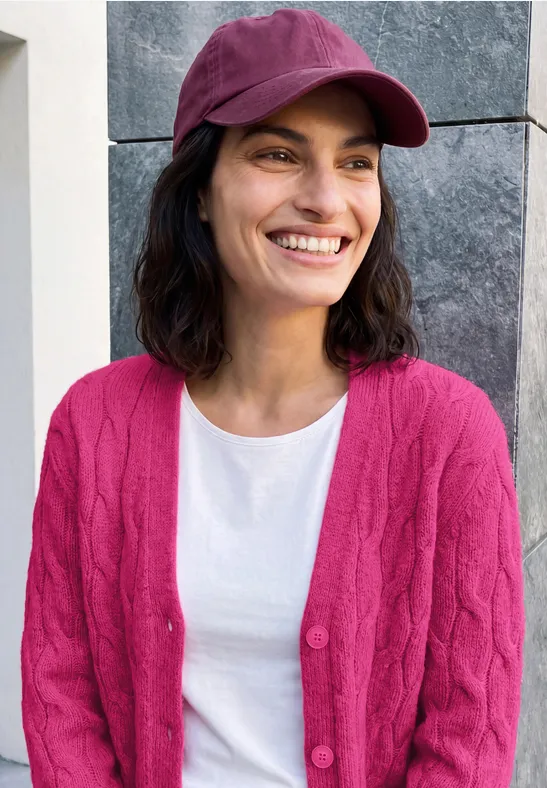 Lächelnde Frau mit dunkelbraunen Haaren, pinker Mütze und Cardigan über weißem T-Shirt vor grauer Wand.
