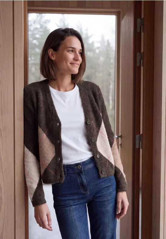 Eine Frau in Jeans, weißem Shirt und kariertem Cardigan lehnt an einem Türrahmen und blickt nach draußen.