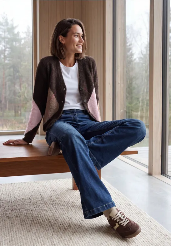 Frau in Jeans und Cardigan sitzt auf Fensterbank und schaut entspannt ins grüne Freie.