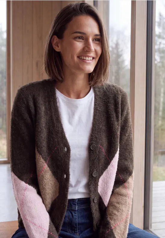 Lächelnde Frau im Strickcardigan, weißem Shirt und Jeans steht vor einem Fenster im hellen Raum.