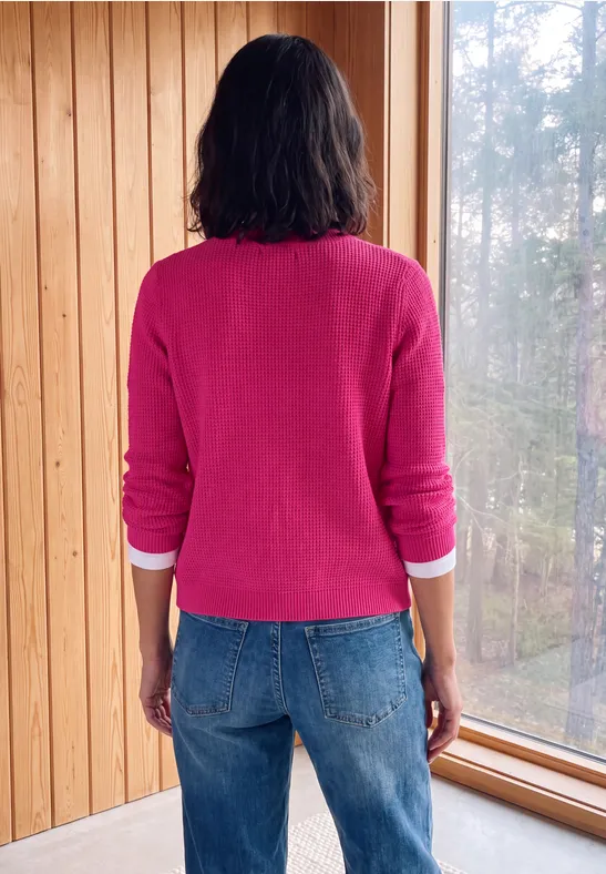 Een persoon in een roze trui en jeans staat met de rug naar de camera bij een houten raam en kijkt naar buiten.