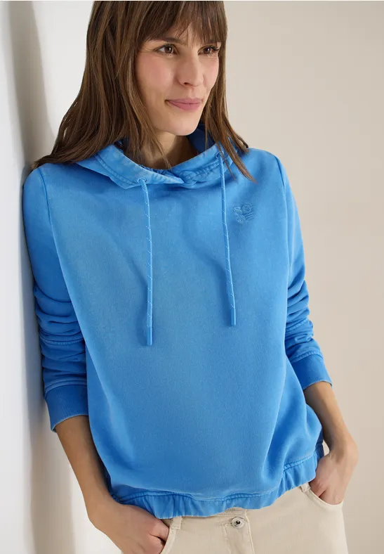Femme en sweat à capuche bleu clair appuyée contre un mur, mains dans les poches.