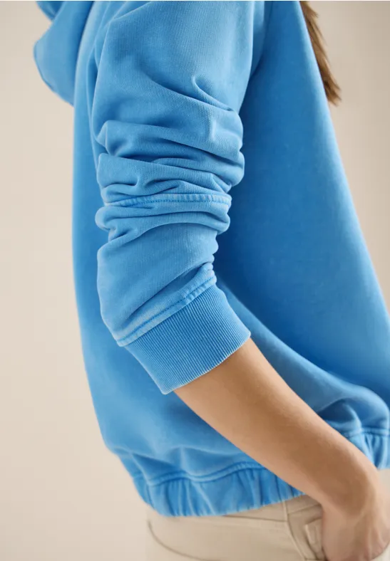Personne en sweat bleu et pantalon beige, vue de côté sur fond clair.