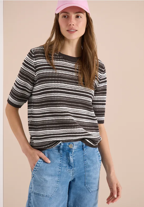 Frau in gestreiftem Schwarz-Weiß-Grau-Shirt und heller Jeans, Hände in den Taschen.