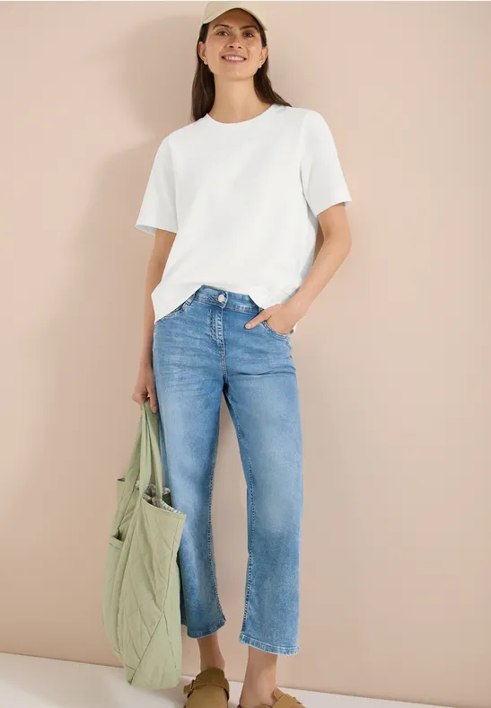 Vrouw met wit T-shirt en lichtblauwe jeans houdt een groene tas vast tegen een beige muur.