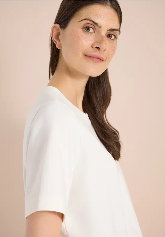 Vrouw in profiel met bruin haar, draagt een eenvoudig wit T-shirt voor een lichte achtergrond.