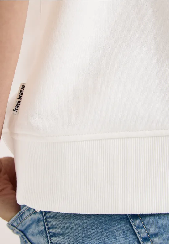 Zoom op de zoom van een witte trui met klein label en blauwe spijkerbroek.